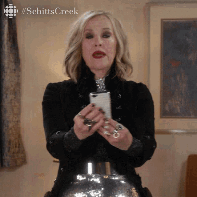 Moira, a woman, from Schitt's Creek (TV show) is holding a cell phone in front of her. Captions "Is it on? Can they see us?"